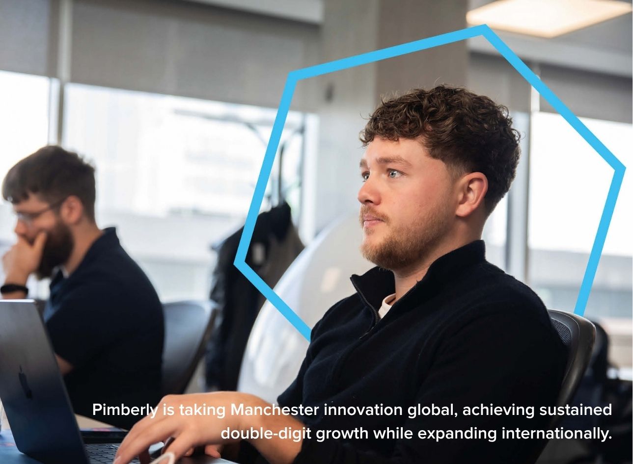 Two people are working in a modern office, using laptops at desks within an open-plan workspace. Large windows provide natural light, while a blue hexagonal graphic overlay frames the foreground subject. The image includes the text: "Pimberly is taking Manchester innovation global, achieving sustained double-digit growth while expanding internationally." The scene highlights digital innovation, business growth, international expansion and the success of a Greater Manchester company in global markets.