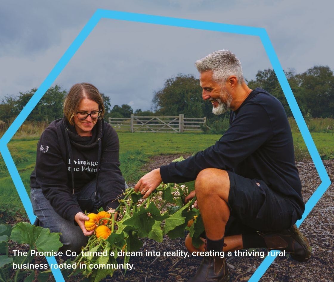 Two people are harvesting yellow squash and other produce in an outdoor growing area at a rural community farm. Raised planting beds, grassland and trees are visible in the background beneath an overcast sky. The image is framed with a blue hexagonal graphic overlay and includes the text: "The Promise Co: turned a dream into reality, creating a thriving rural business rooted in community." The scene highlights sustainable entrepreneurship, local food growing, community impact and rural business success.